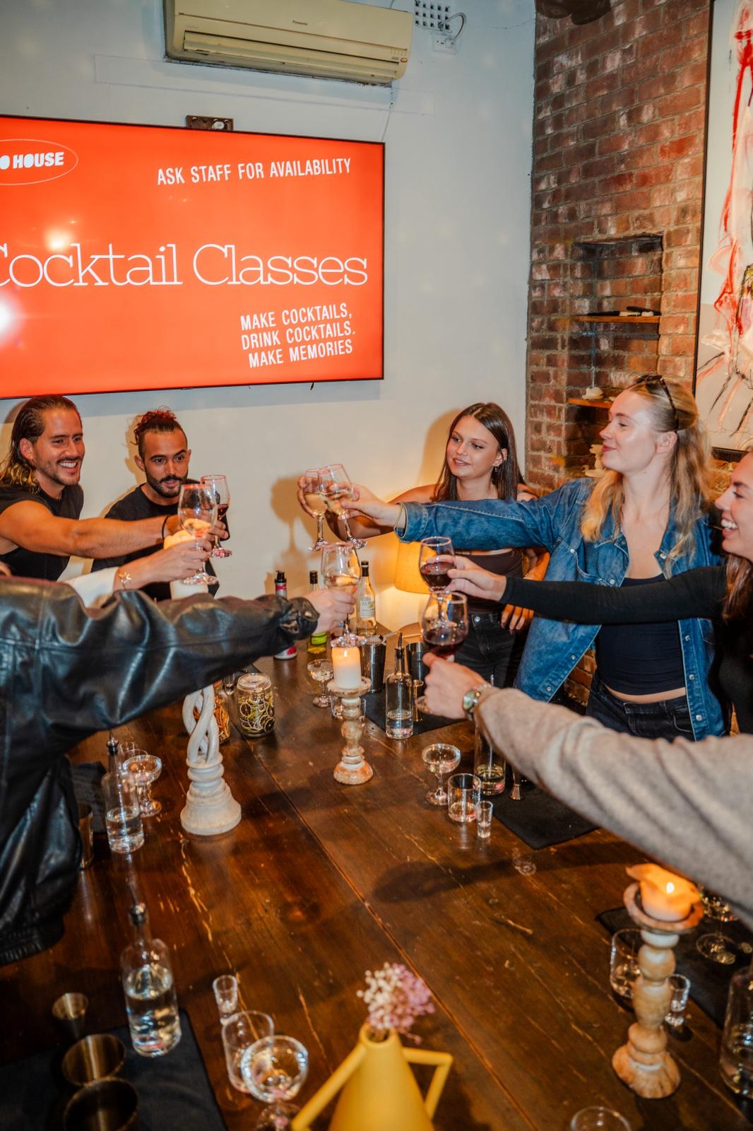 Group of friends clinking cocktail glasses around a wooden table in a cozy room.