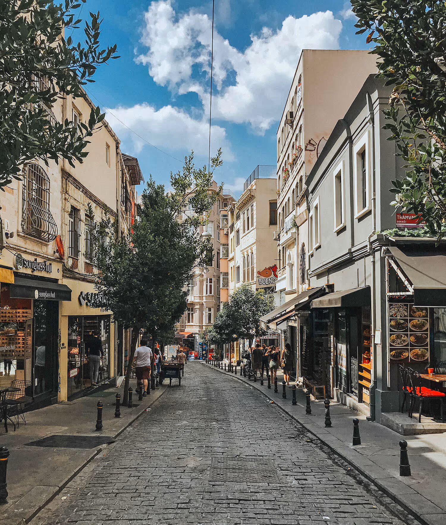 Cobblestone street lined with shops and cafes under a blue sky with scattered clouds, featuring pedestrians walking and trees along the sidewalks.