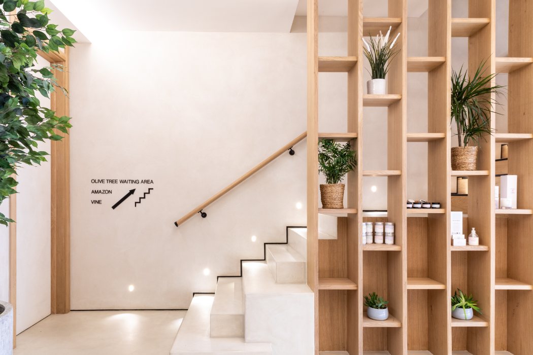 Minimalist interior with light-colored stairs, wooden handrail, wooden shelving unit with potted plants and skincare products, and directional text on the wall.