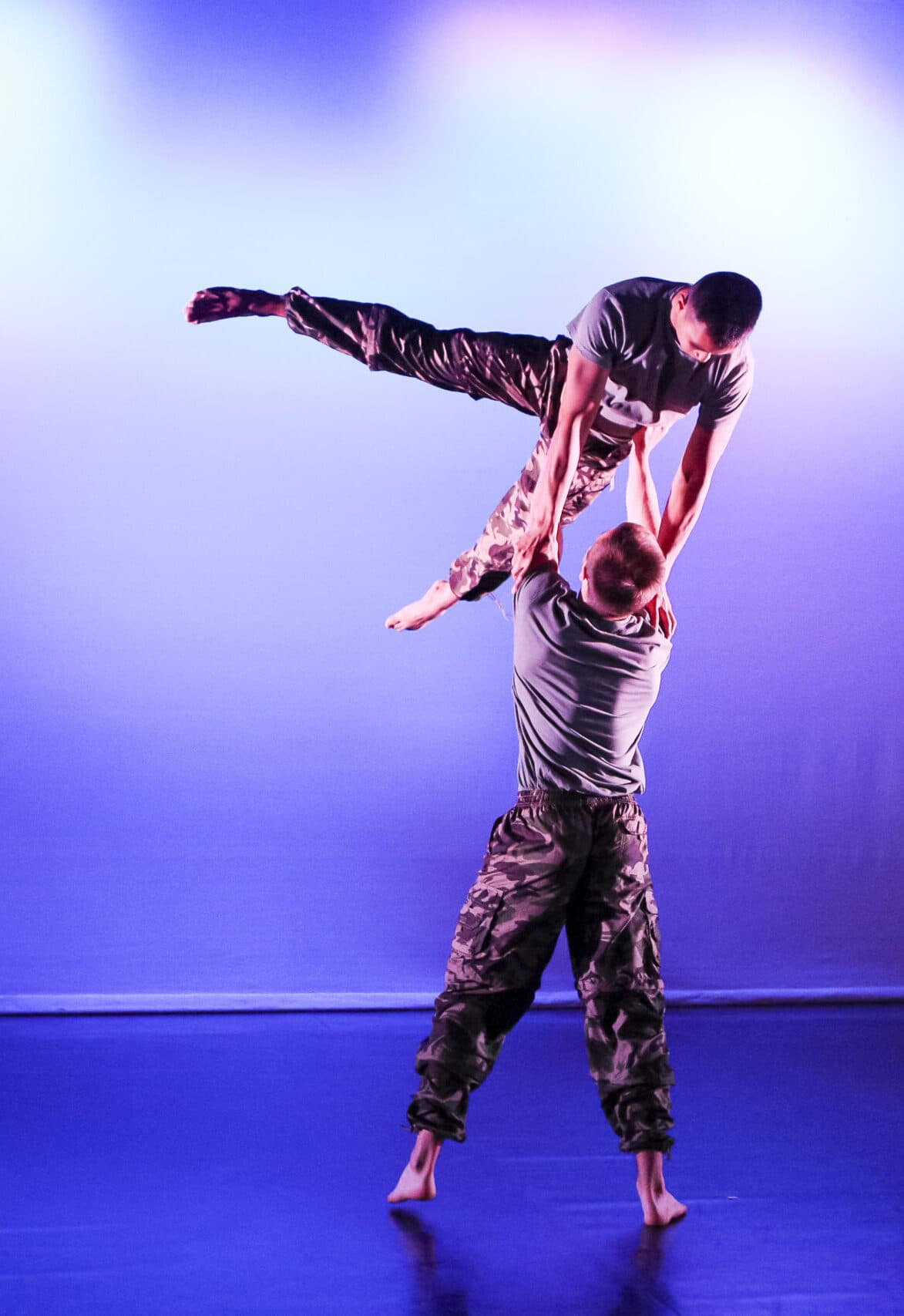 Two men in camouflage pants and gray shirts performing an acrobatic lift on stage with a blue-purple gradient background.