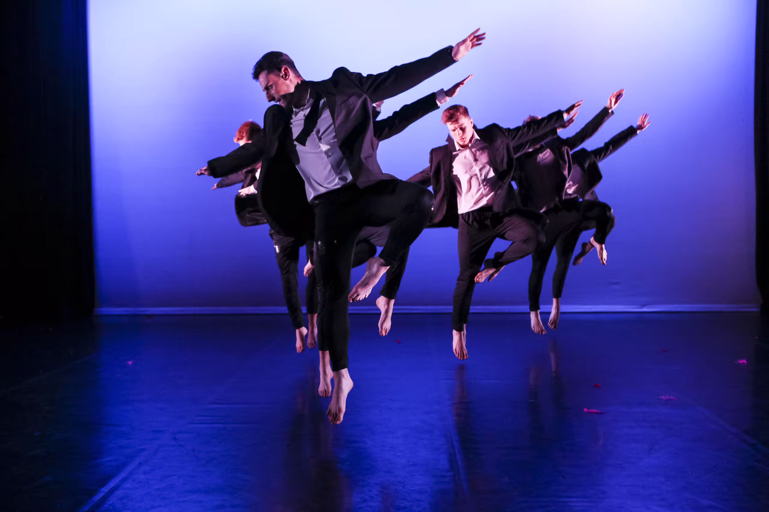 Group of male dancers in black suits jumping with arms outstretched on a blue-lit stage.