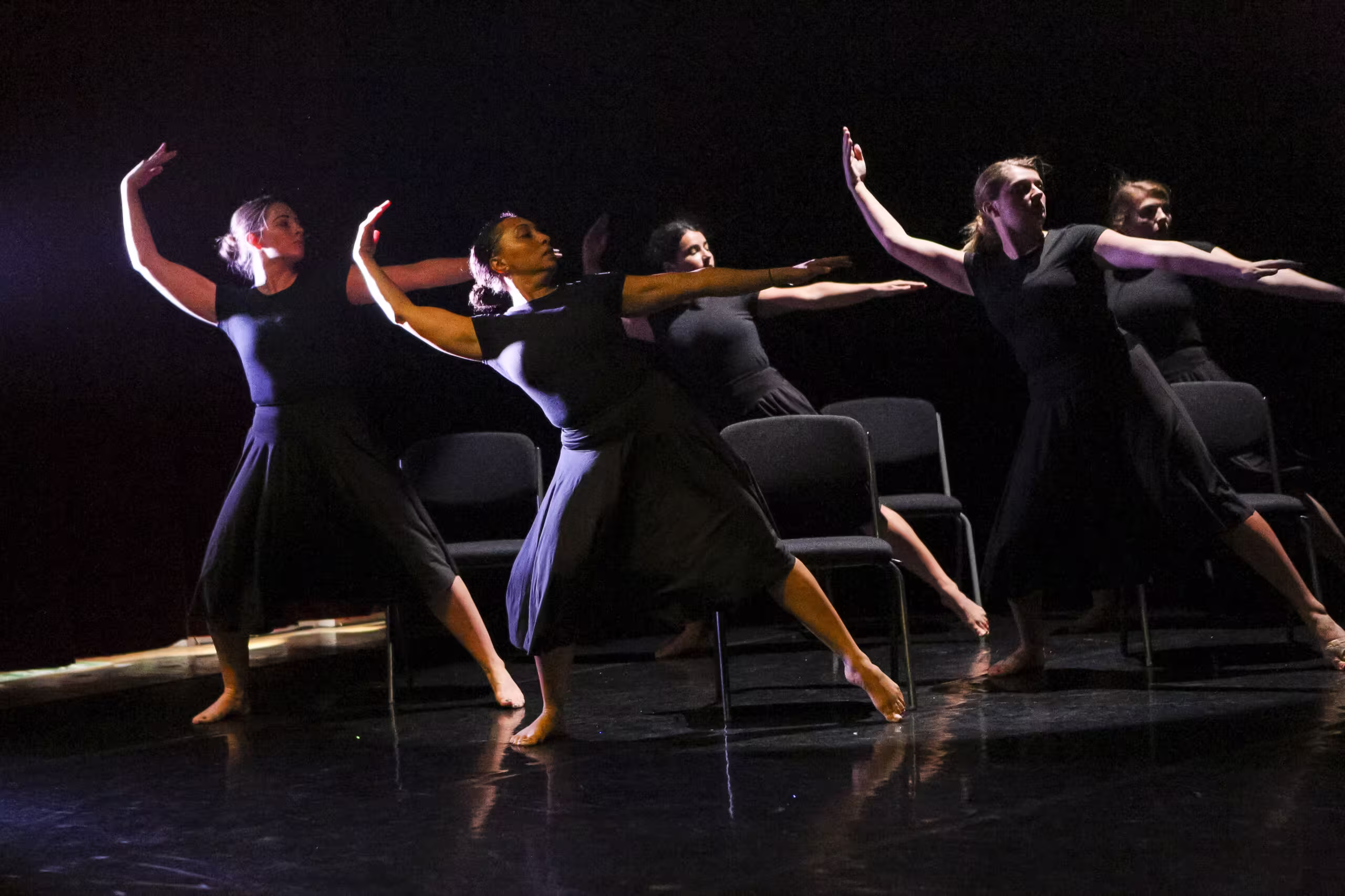 Five dancers in black dresses performing a synchronized contemporary dance with chairs on a dark stage.