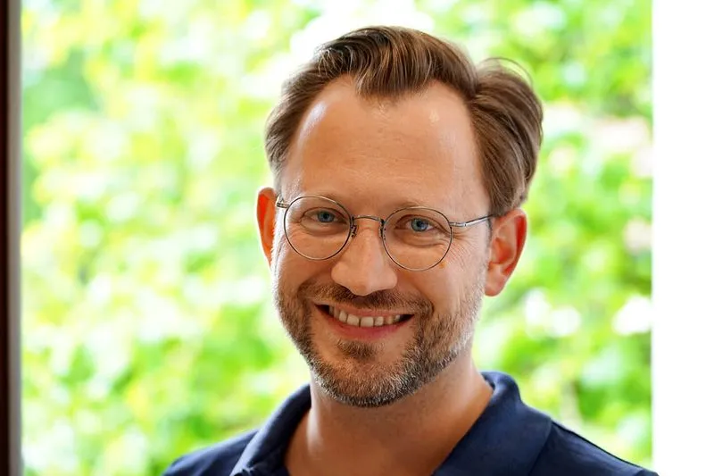 Portrait Prof. Dr. med. Ulrich Straßen, mit runder Brille und Bart, lächelnd vor unscharfem grünen Hintergrund