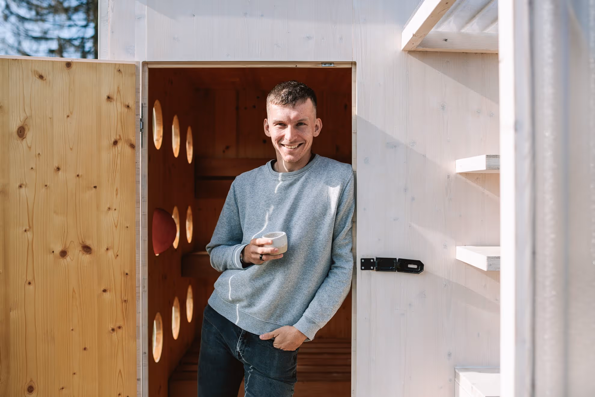 Ivan Zaťko, stojící ve dveřích finské sauny, drží hrnek s kávou a usmívá se.