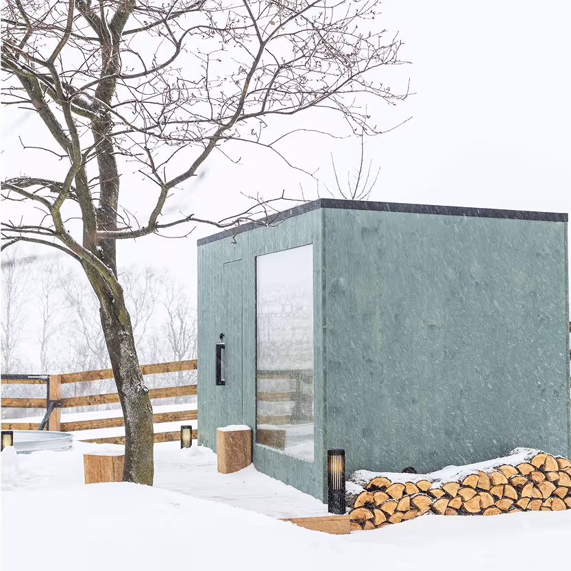 Designová venkovní sauna v zasněžené zahradě s připravenou zásobou palivového dřeva vyskládanou podél boční stěny.