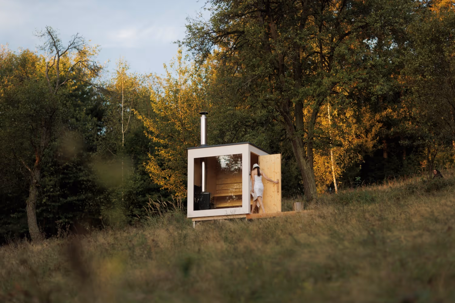 Outdoor Finnish sauna with a large window and a wood-burning stove, standing in a meadow by a forest. A woman in a white sheet and a sauna hat is entering the sauna.