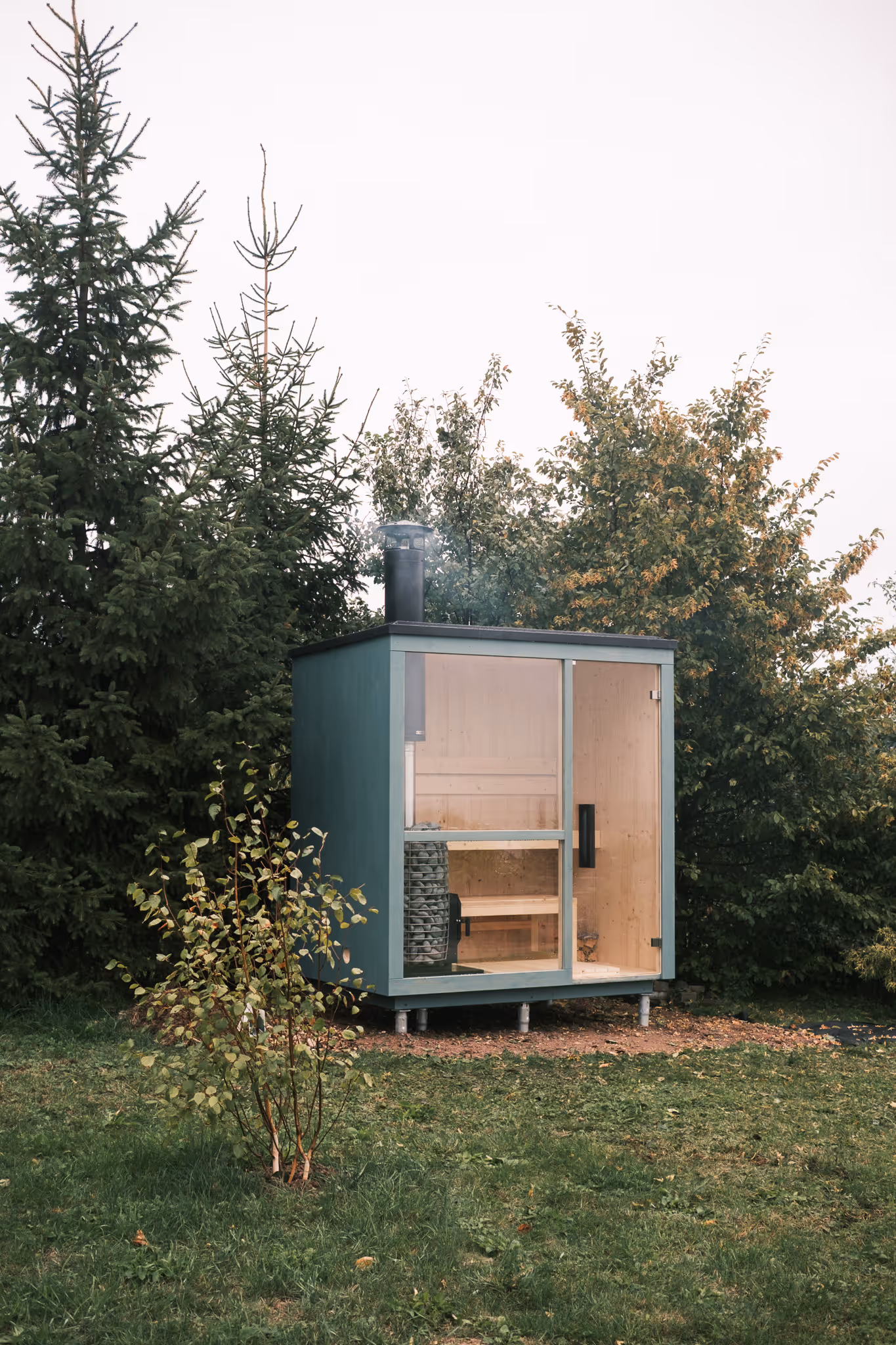 Venkovní finská sauna v odstínu Dusty Teal s velkým oknem a skleněnými dveřmi, umístěná na kraji zahrady se stromy v pozadí.
