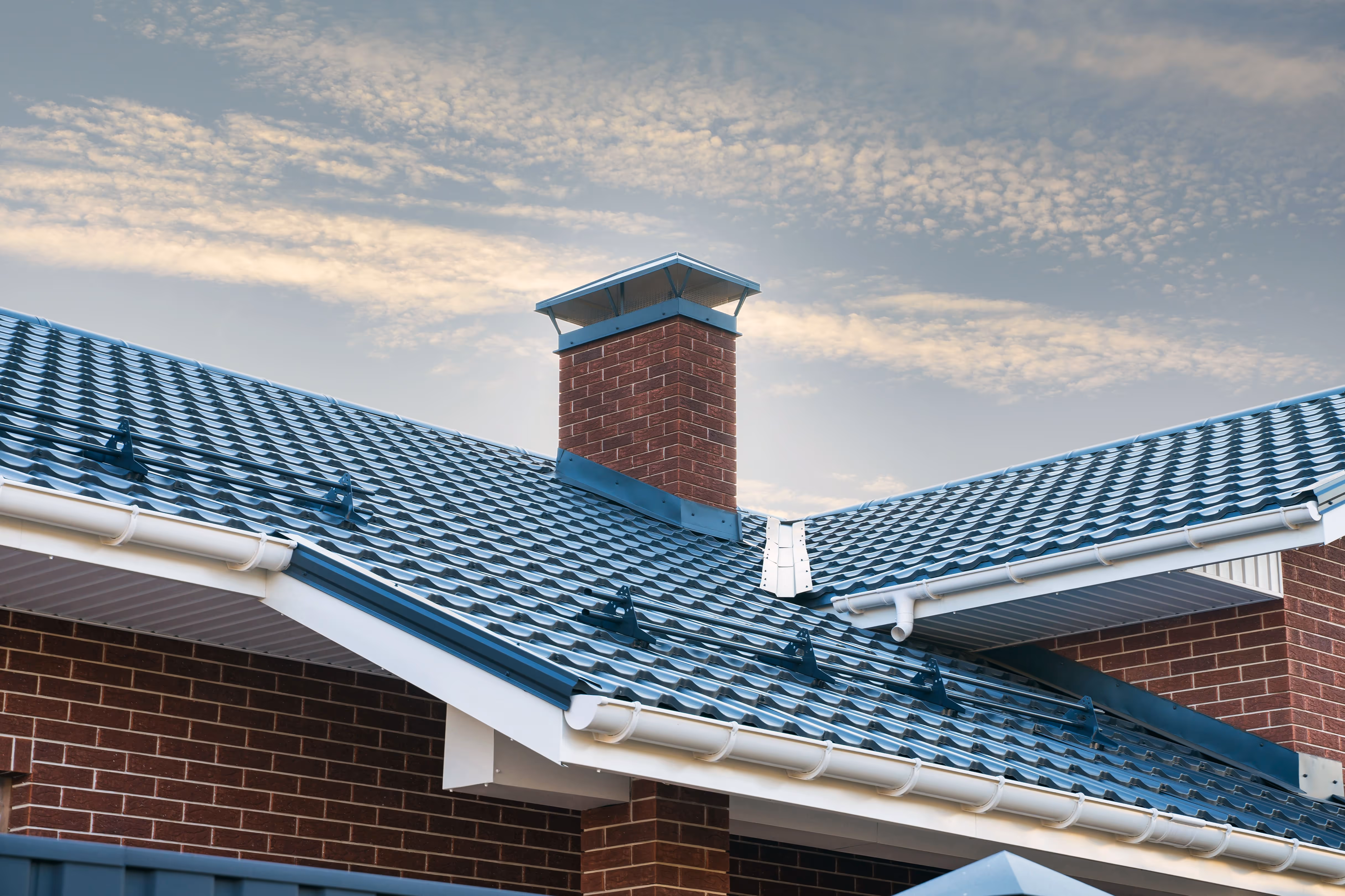Blue metal tile roof with a brick chimney and white gutters under a partly cloudy sky.