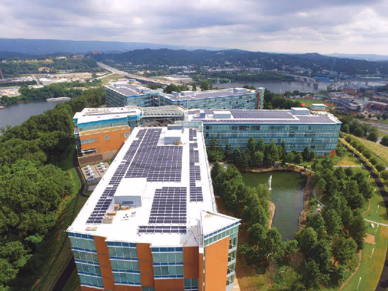 Aerial view of a modern office complex with glass and brick buildings, solar panels on rooftops, surrounding trees, a water fountain in a pond, and a river in the background.