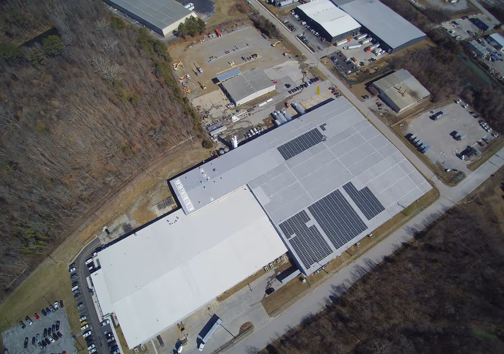 Aerial view of an industrial complex with large white-roofed buildings, some equipped with solar panels, surrounded by parking lots, roads, and wooded areas.