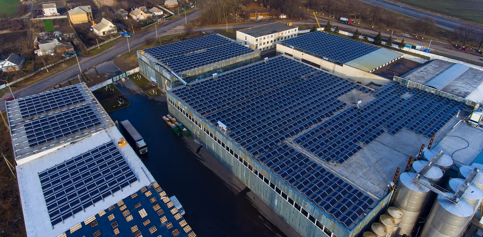 Aerial view of multiple industrial buildings with large solar panel arrays on their roofs and surrounding silos and pallets.