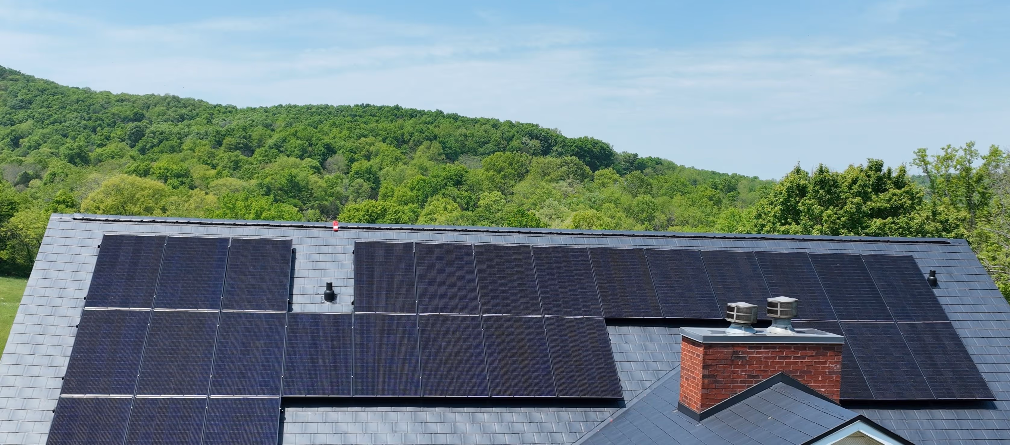 Roof with multiple solar panels installed against a backdrop of green trees and blue sky.