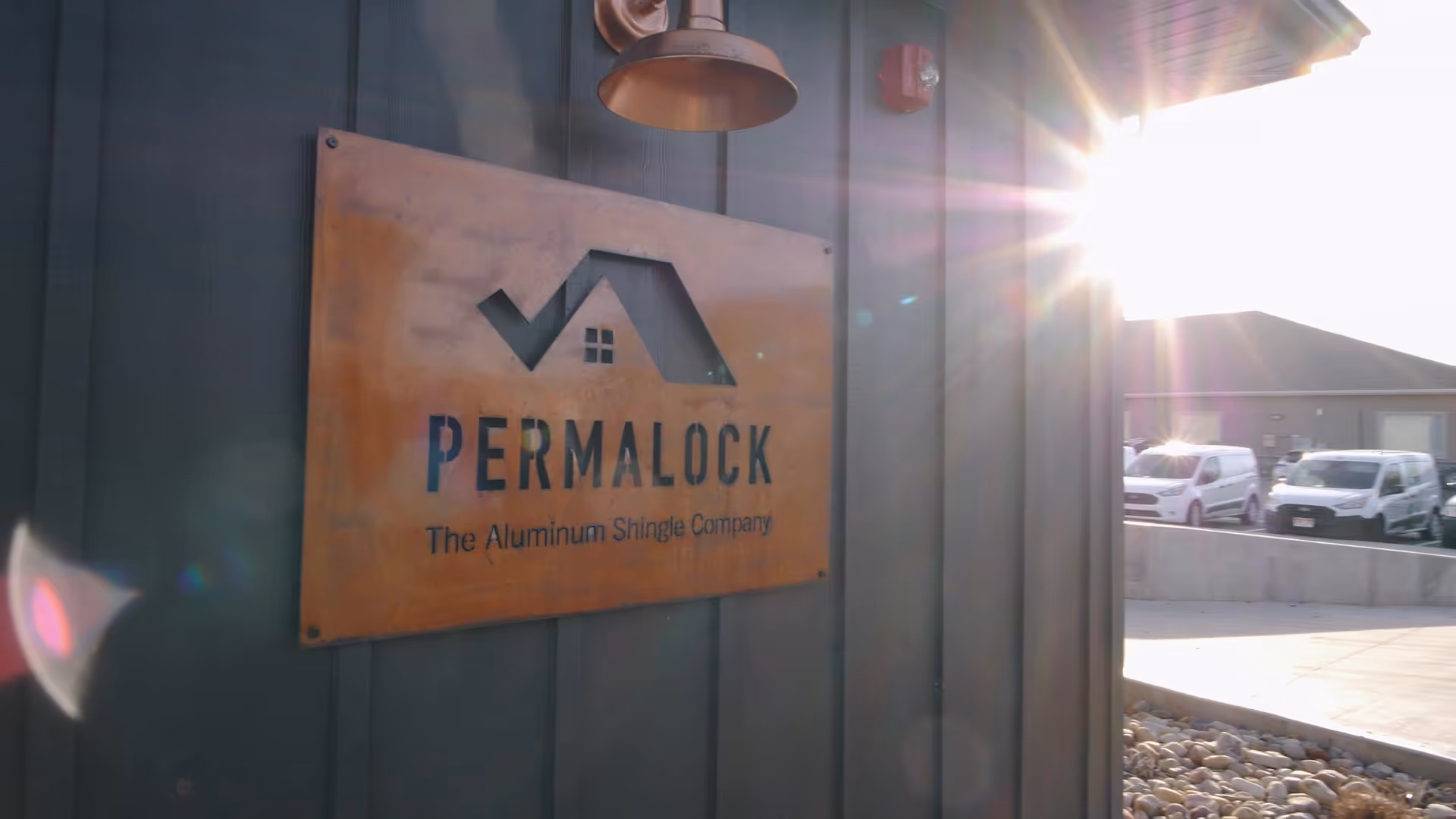 Rust-colored metal sign for Permalock, The Aluminum Shingle Company, mounted on a dark vertical panel wall with sunlight flare.
