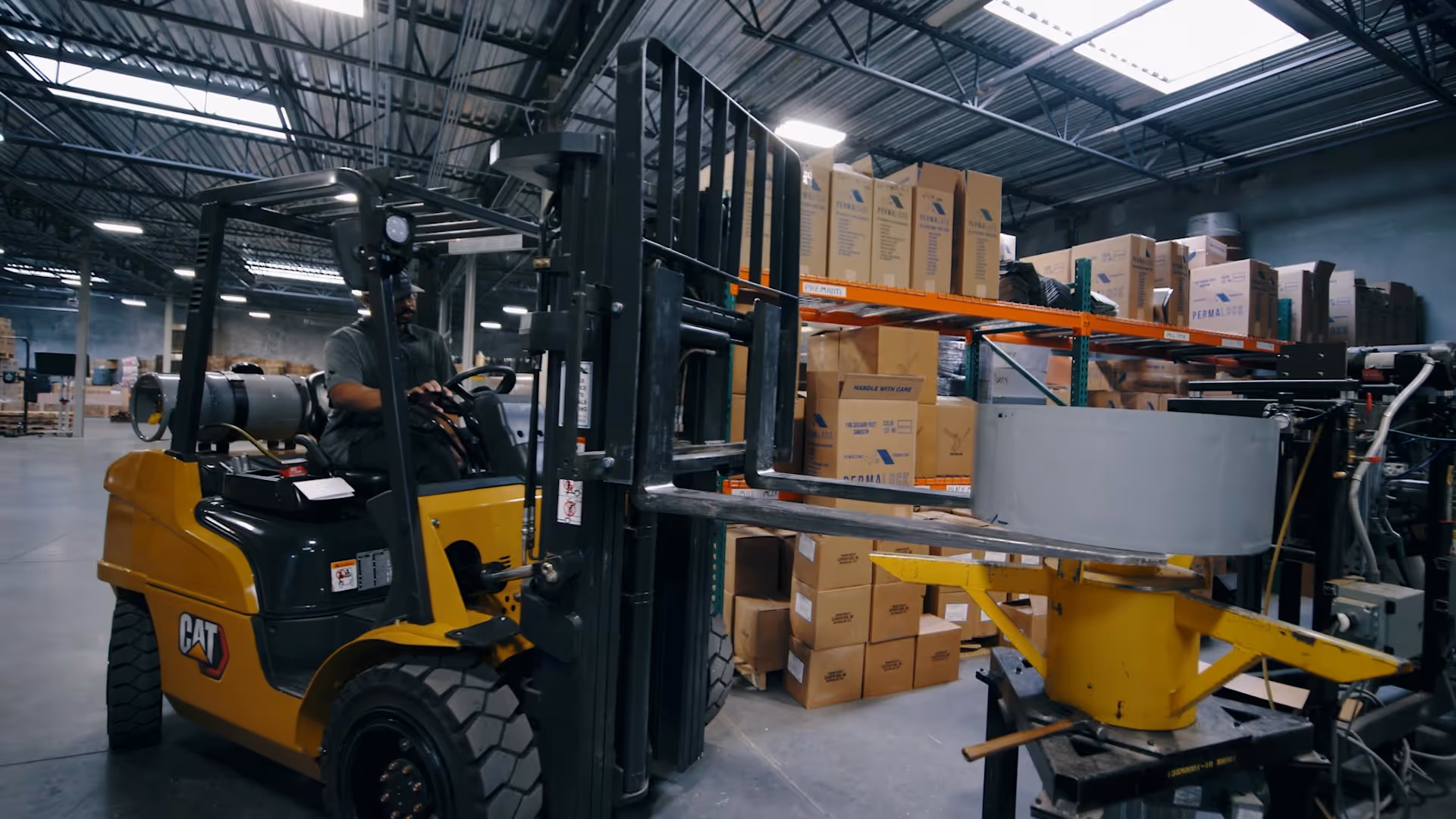 Worker operating yellow CAT forklift in a warehouse near stacked cardboard boxes and industrial equipment.