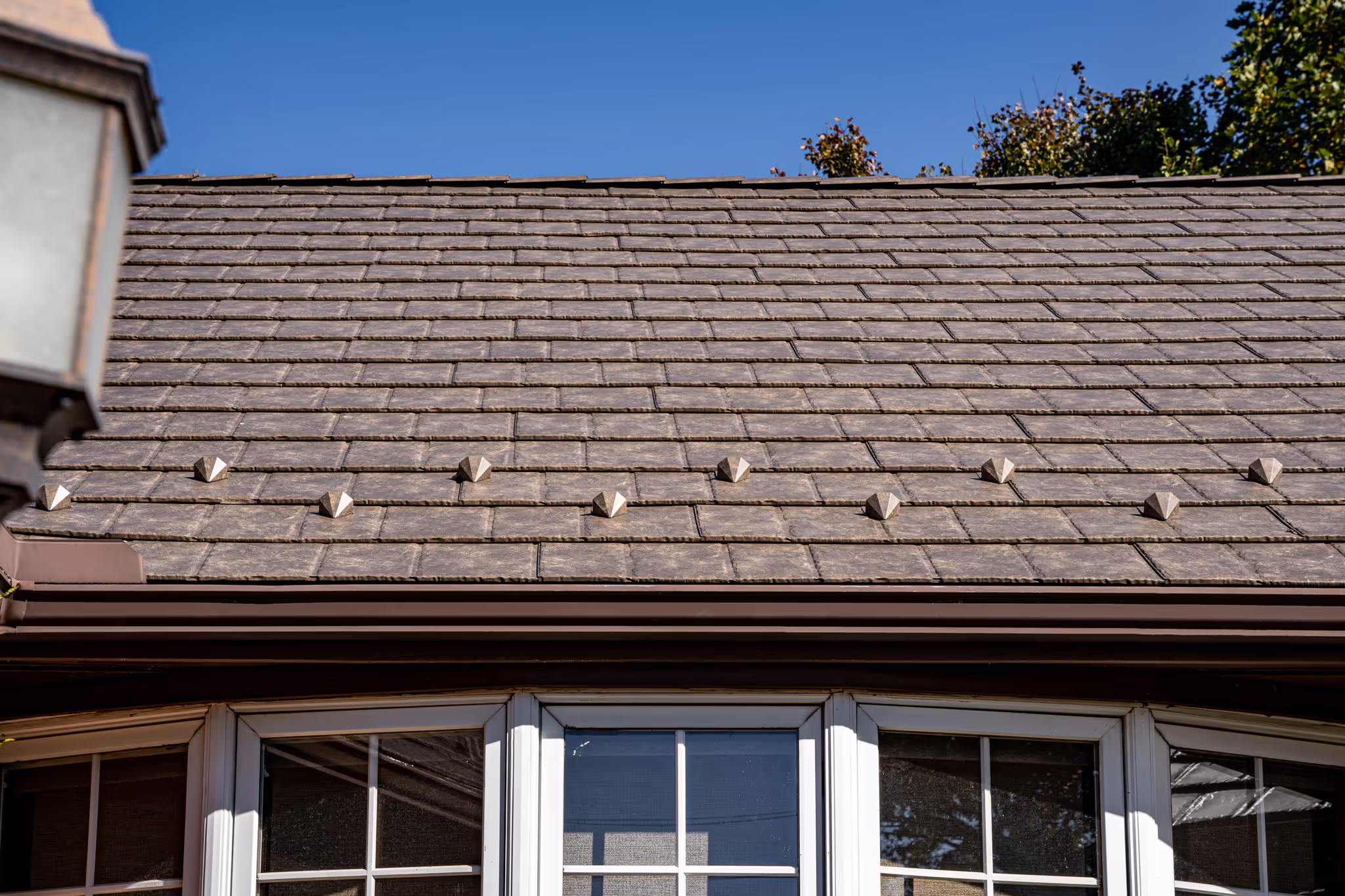 Close-up of a brown tiled roof with decorative metal spikes and white-framed windows below under a clear blue sky.
