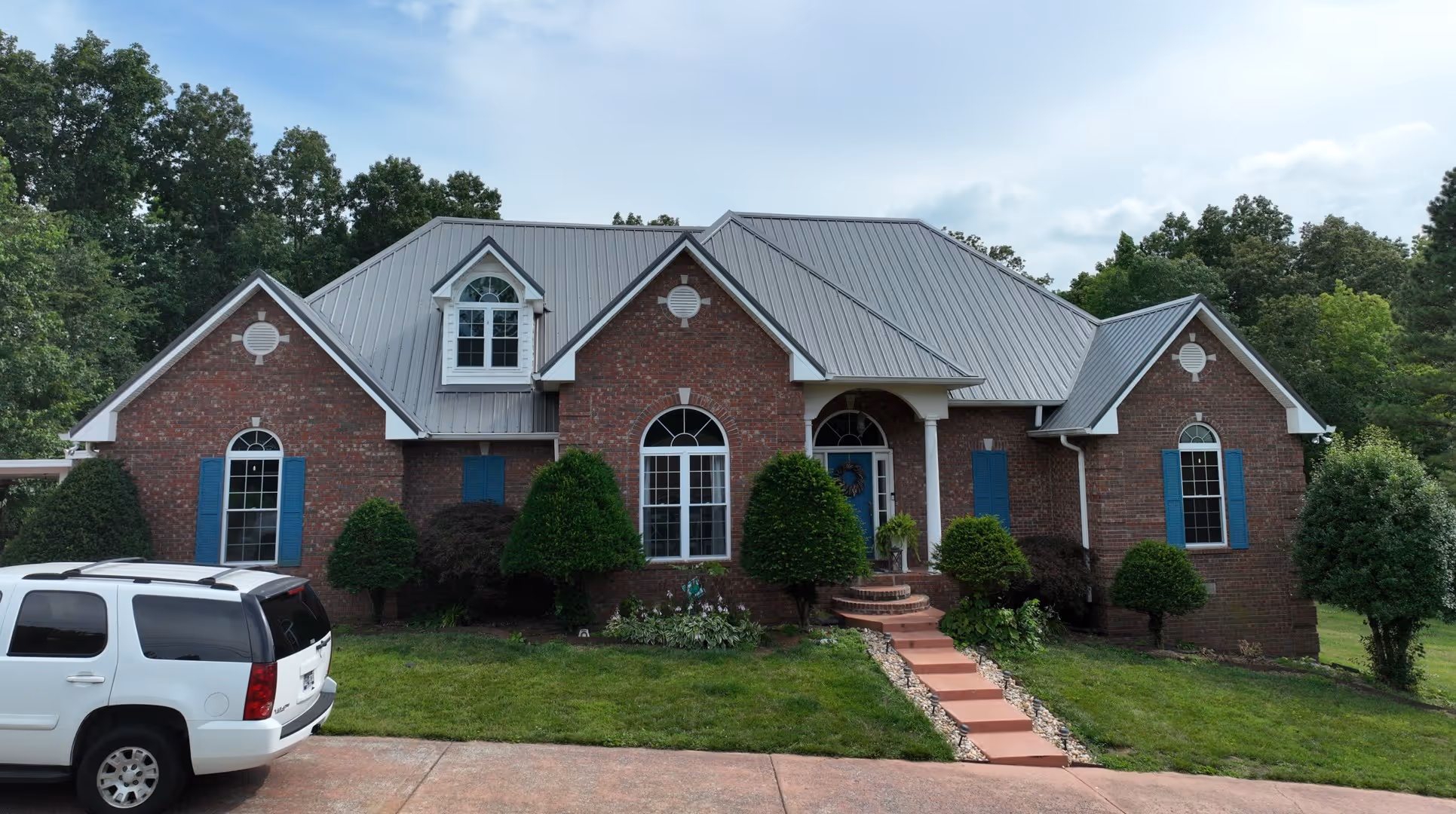 Brick house with gray metal roof, blue shutters, arched windows, a blue front door, manicured bushes, and a white SUV parked in the driveway.