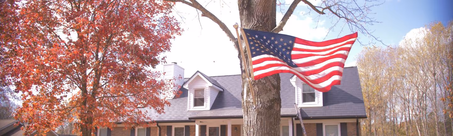 American flag waving on a pole attached to a tree in front of a house with autumn foliage.