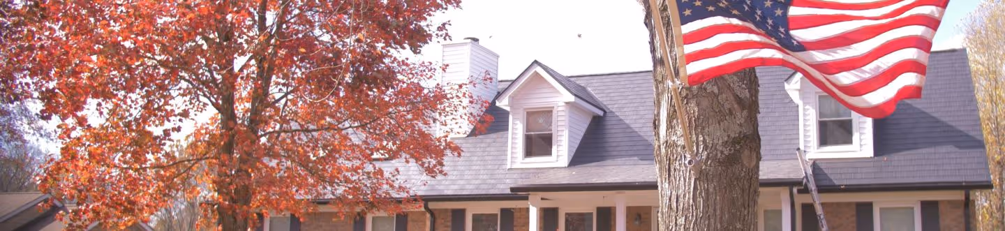 American flag flying on a tree trunk in front of a house with autumn-colored leaves on nearby trees.