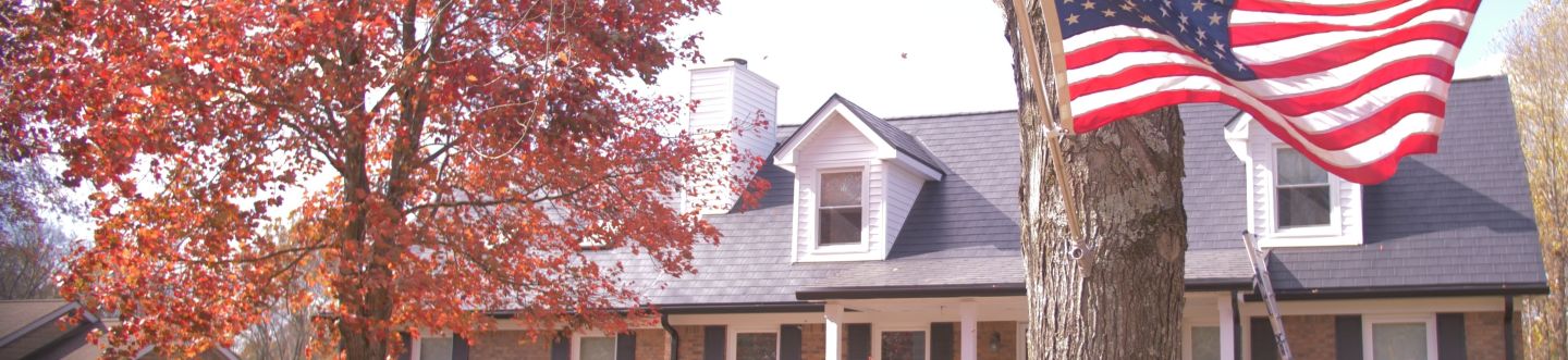 American flag flying on a tree trunk in front of a house with autumn-colored leaves on nearby trees.