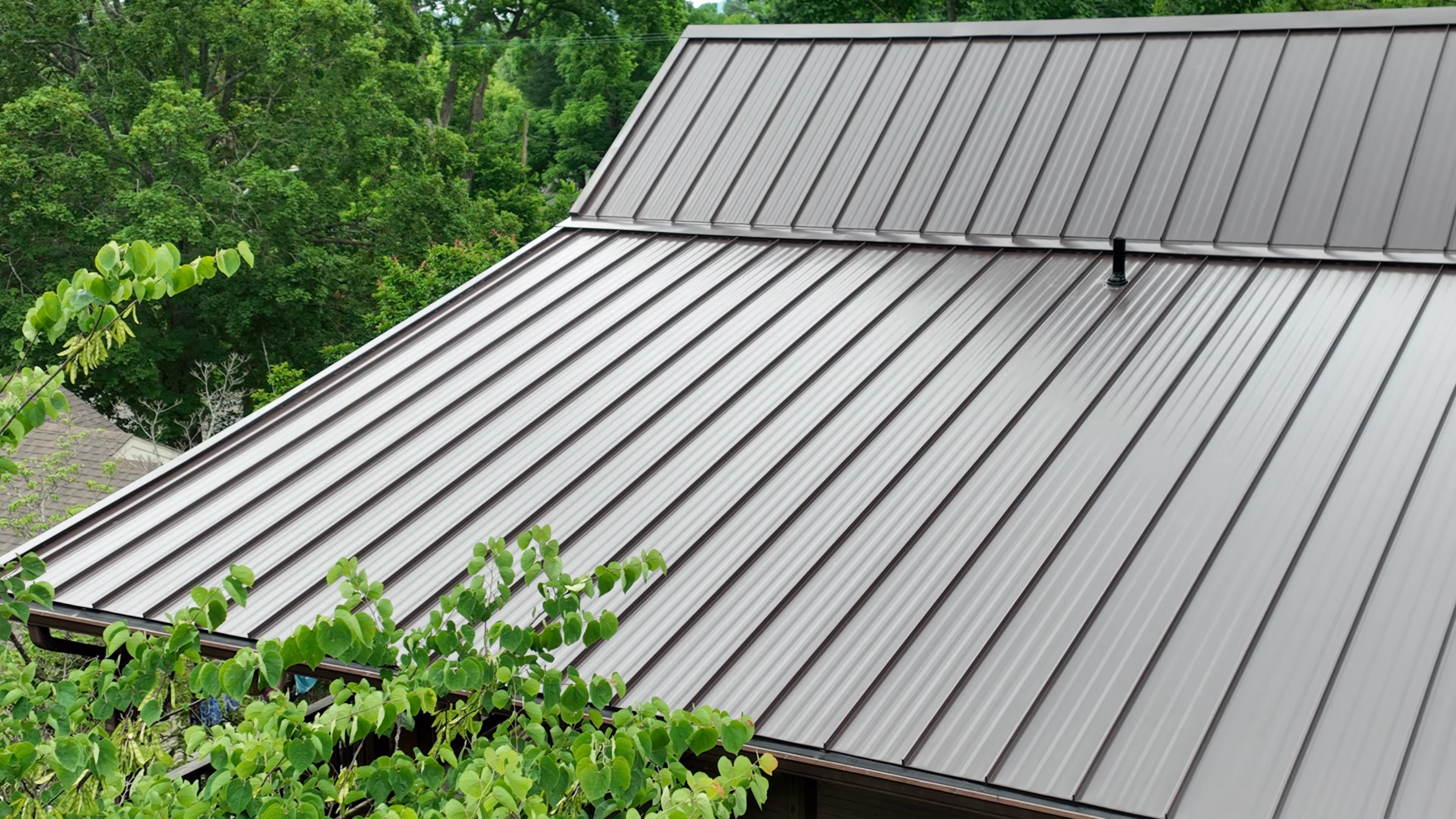 Close-up of a gray metal roof with vertical ridges surrounded by green leafy trees.