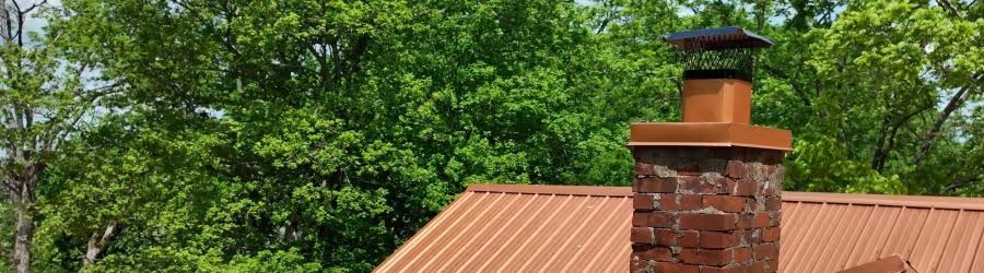 Brick chimney with metal cap on a rust-colored metal roof, surrounded by green leafy trees.