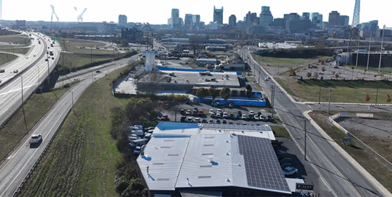 Aerial view of an industrial area with buildings, parking lots, and adjacent highways leading toward a distant city skyline under clear skies.
