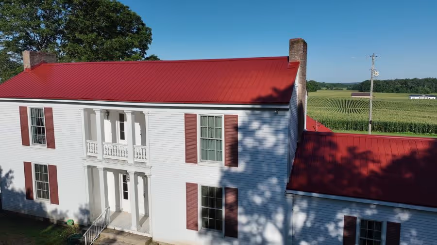 Two-story white farmhouse with red metal roof and shutters next to a green field under a clear blue sky.