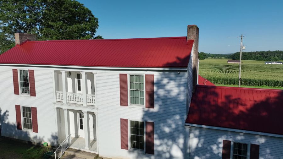 Two-story white farmhouse with red metal roof and shutters next to a green field under a clear blue sky.