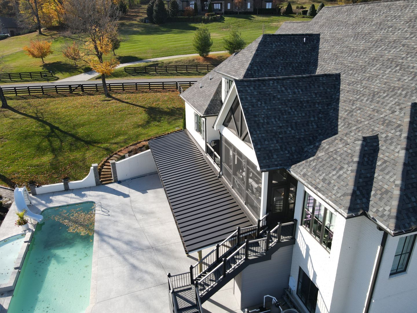 Aerial view of a large white house with gray shingle roof, black metal patio awning, outdoor stairs, and a swimming pool with a slide surrounded by autumn trees and green lawn.