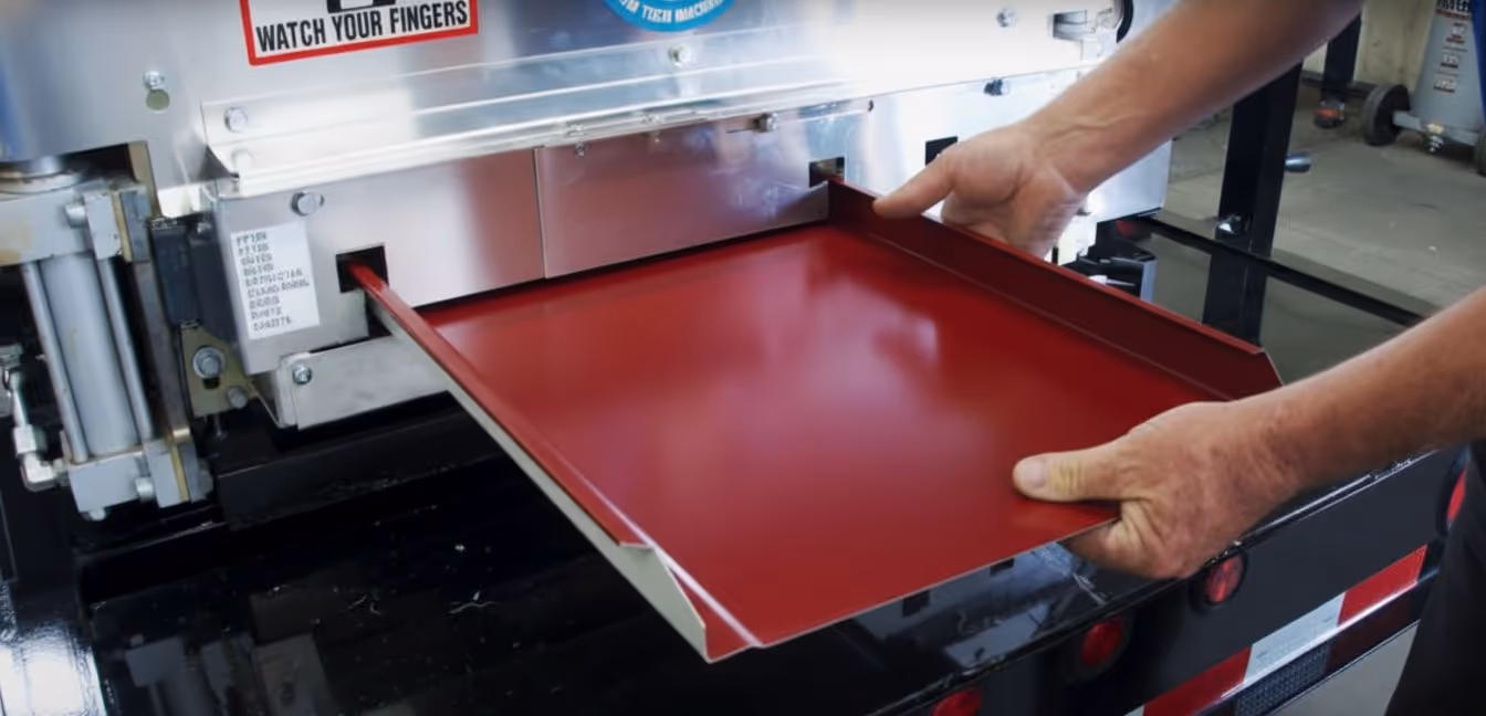 Hands operating a metal bending machine shaping a red sheet of metal with a caution sign reading 'WATCH YOUR FINGERS'.