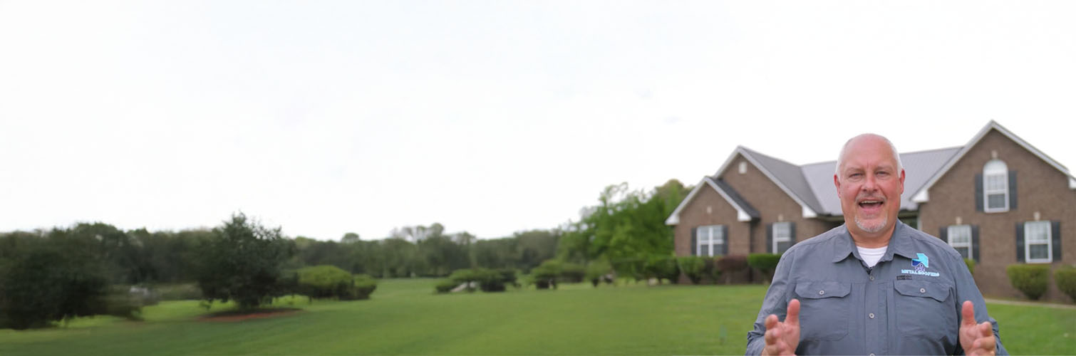 Man in a gray work shirt standing on a lawn with a brick house and trees in the background.