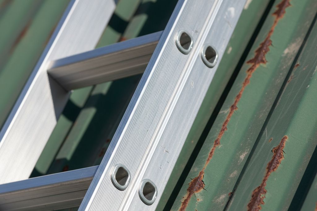Close-up of a silver aluminum ladder leaning against a green corrugated metal surface with rust streaks.