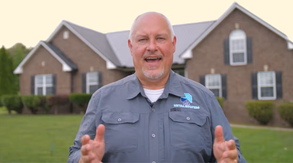 Smiling middle-aged man with short gray hair and goatee, wearing a gray button-up shirt, standing in front of a large brick house with a green lawn.