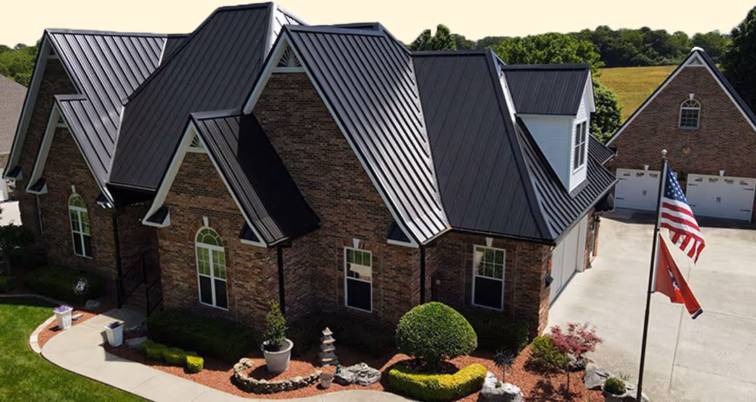 Large brick house with steep black metal roof, manicured bushes, and an American flag on a driveway beside a detached garage.
