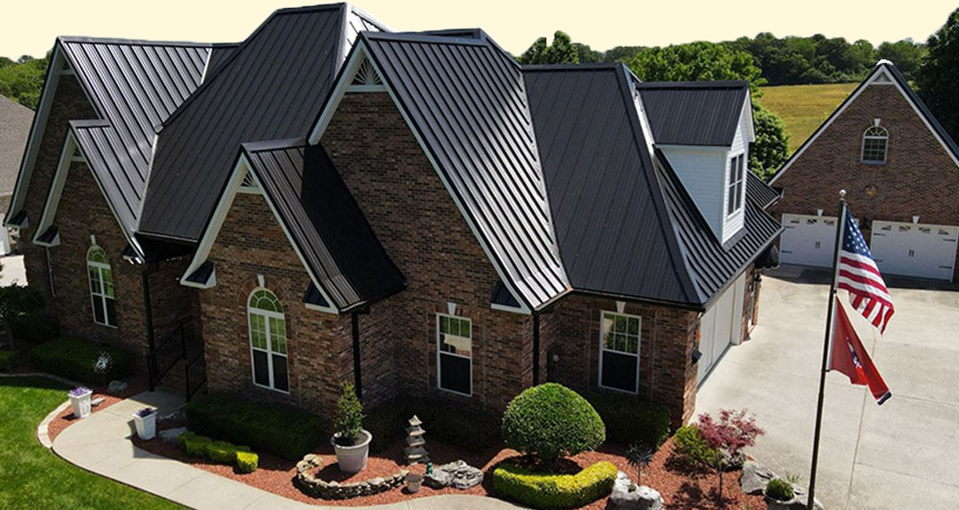 Large brick house with steep black metal roof, manicured bushes, and an American flag on a driveway beside a detached garage.