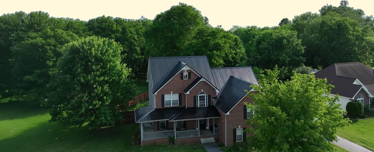 Two-story brick house with black metal roof surrounded by green trees and lawn.