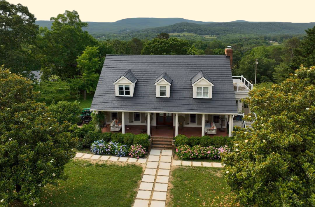 Cottage-style house with a gray roof, front porch with swing chairs, flower beds, and green lawn surrounded by trees and hills in the background.