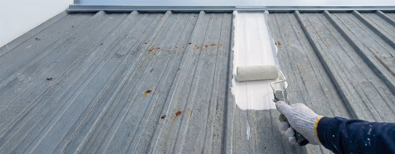 Person wearing a glove painting a rusty metal roof white with a paint roller.