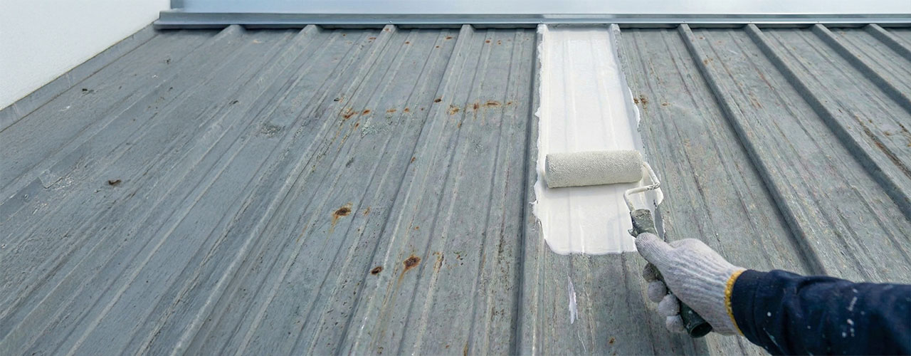 Person wearing a glove painting a rusty metal roof white with a paint roller.