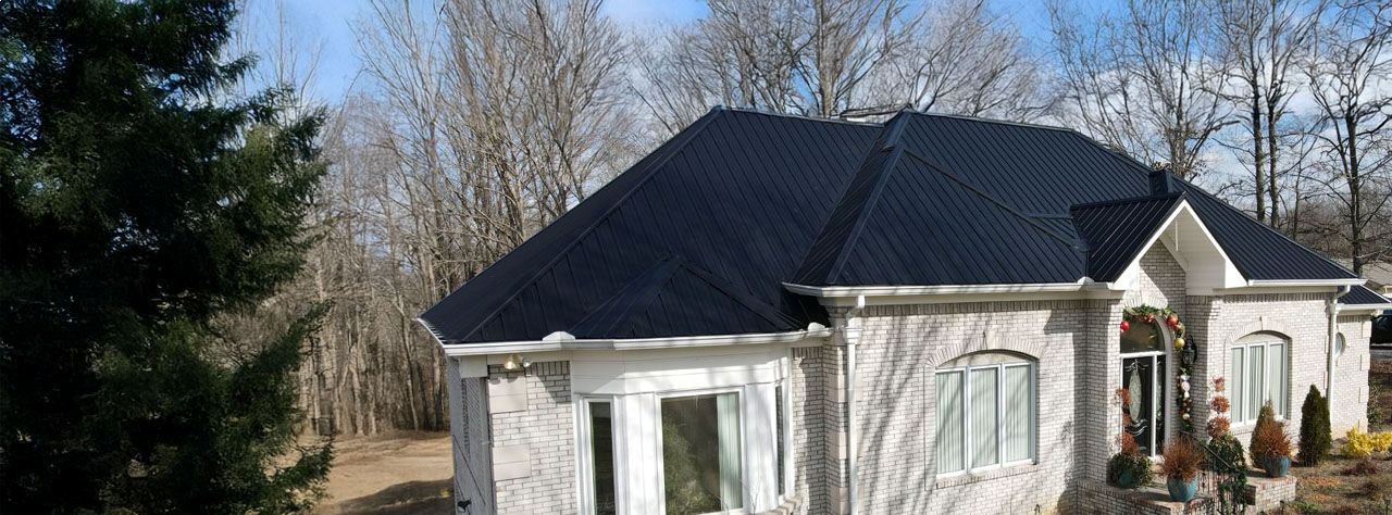 Light gray brick house with a steep black metal roof surrounded by leafless trees and evergreen shrubs.