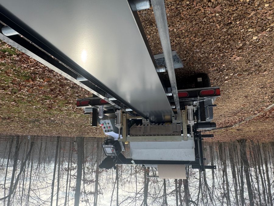 Large industrial machine on a trailer producing a smooth, flat, dark metal sheet with trees and fallen leaves in the background.