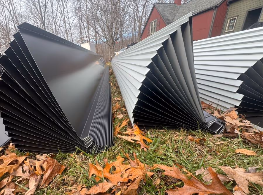 Stacks of black and light gray metal siding panels lying on grass with fallen autumn leaves near houses and bare trees.