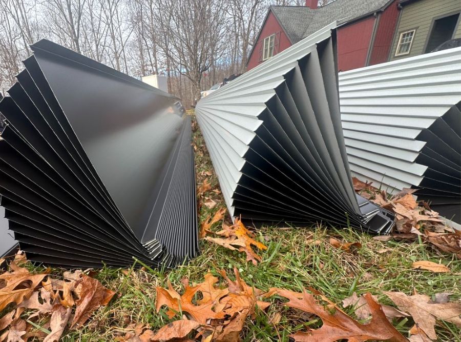Stacks of black and light gray metal siding panels lying on grass with fallen autumn leaves near houses and bare trees.