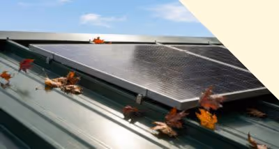 Solar panel installed on a green metal roof with scattered autumn leaves around it under a blue sky with clouds.