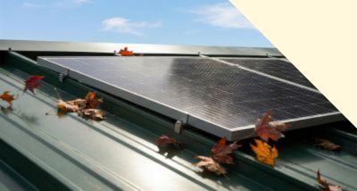 Solar panel installed on a green metal roof with scattered autumn leaves around it under a blue sky with clouds.