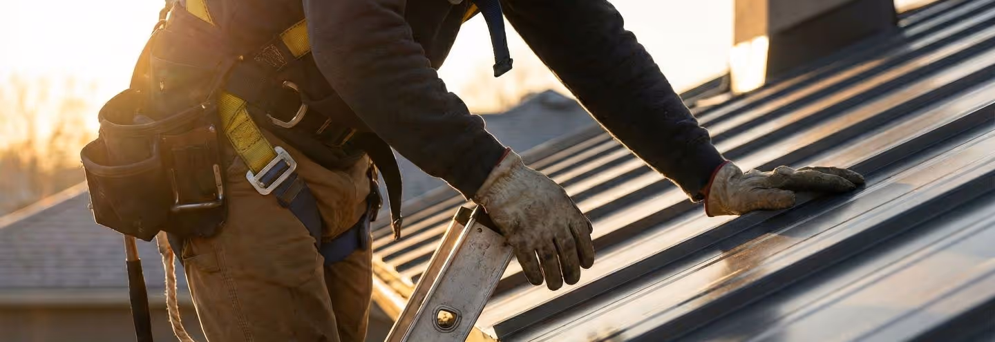 Construction worker wearing gloves and a safety harness working on a metal roof at sunset.