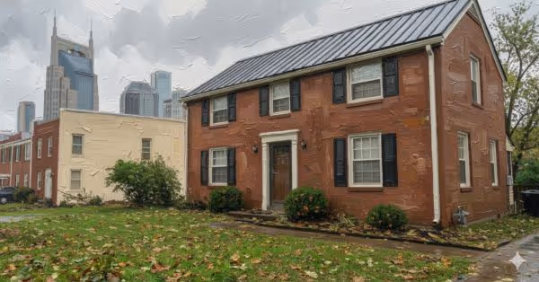 Two-story red brick house with black shutters and white-trimmed windows, set on a lawn with fallen leaves and city skyline in the background.