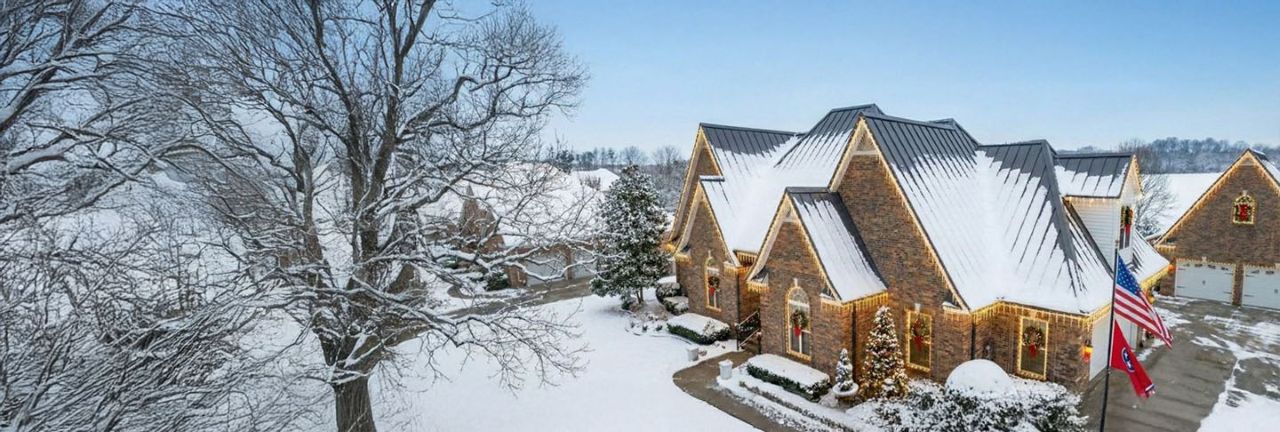 Snow-covered brick house decorated with Christmas wreaths and lights, surrounded by leafless trees and a snowy yard.