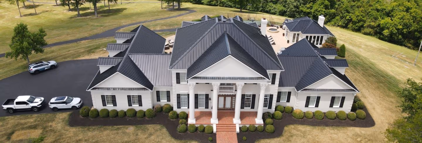 Aerial view of a large white colonial-style house with multiple black metal roofs, tall white columns at the entrance, surrounded by trimmed bushes and a driveway with three parked vehicles.