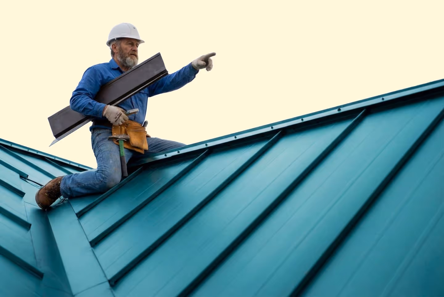 Bearded construction worker wearing a white helmet and blue shirt kneeling on a teal metal roof, holding roofing material and pointing forward.